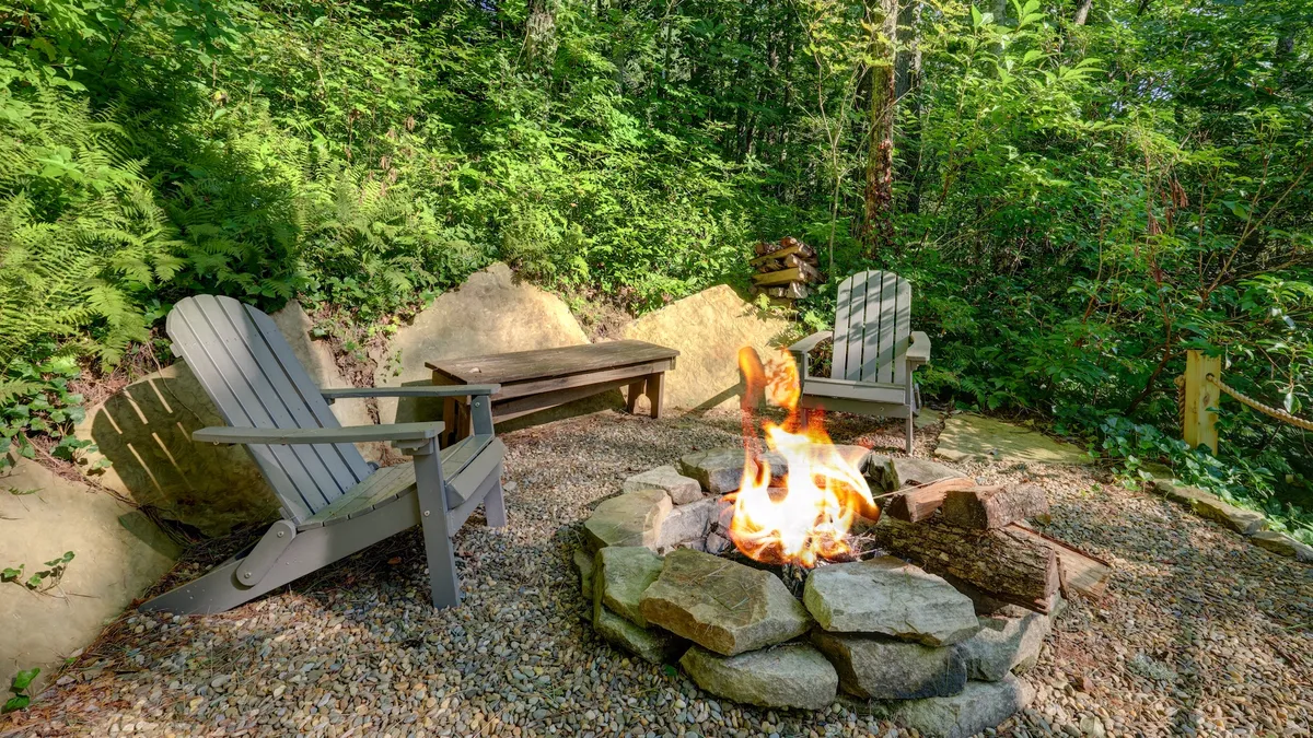 A cozy outdoor fire pit is encircled by Adirondack chairs, set amidst lush greenery. The arrangement suggests a welcoming space for gathering, storytelling, and stargazing, with a backdrop of tranquil nature.