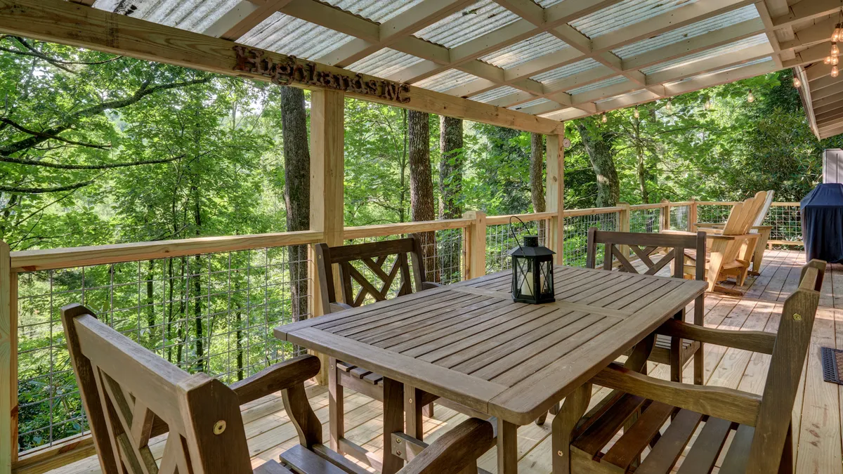 A serene deck with a wooden dining table overlooks surrounding trees, accented by an outdoor lantern and rustic deck railing. The setting is ideal for morning coffee, family gatherings, or intimate meals amid the beauty of nature, with the promise of sunset views.