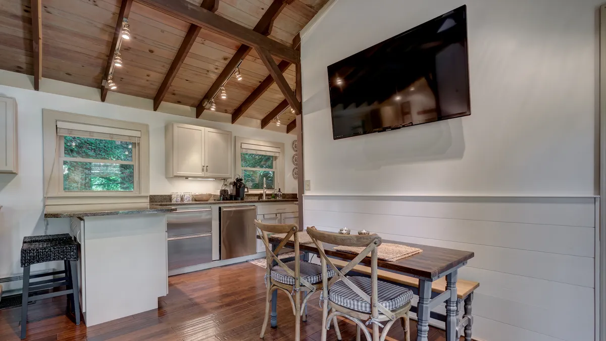 A warm, open kitchen and dining area with a prominent kitchen island, a wooden dining table, exposed wooden beams overhead, and a TV in the background. The space is filled with ambient lighting and welcoming touches, inviting gatherings and culinary activity.