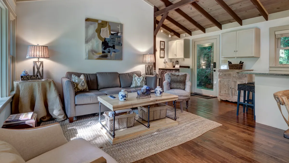 This image showcases a beautifully furnished living room with a welcoming sofa, a coffee table, tasteful artwork, and a peek of the open kitchen area. Large windows allow natural light to fill the space, accentuating the airy and modern atmosphere. The seating arrangement encourages socializing, relaxing, and enjoying the tranquil forest views beyond.