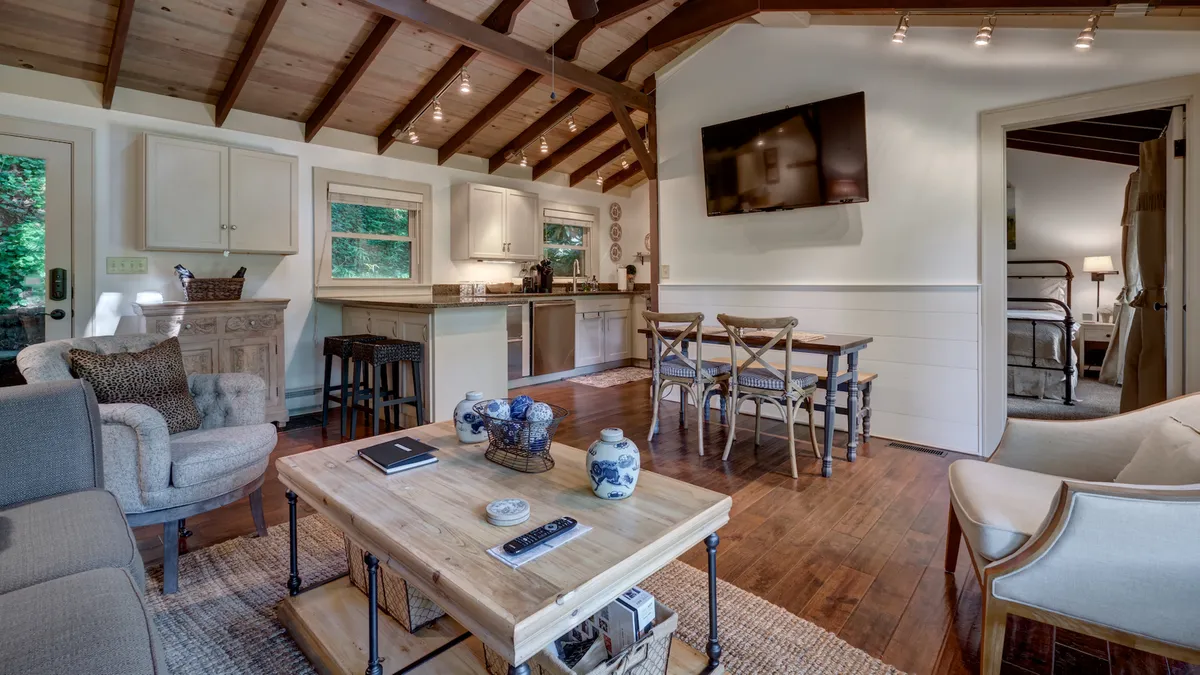 A spacious and light-filled main gathering area featuring exposed wooden beams, a cozy sofa, a wooden dining table set for meals, and a TV. Decorative vases and thoughtful decor elements create a welcoming, modern ambiance. Large windows hint at the greenery outside, further blending indoor elegance with a tranquil natural setting.