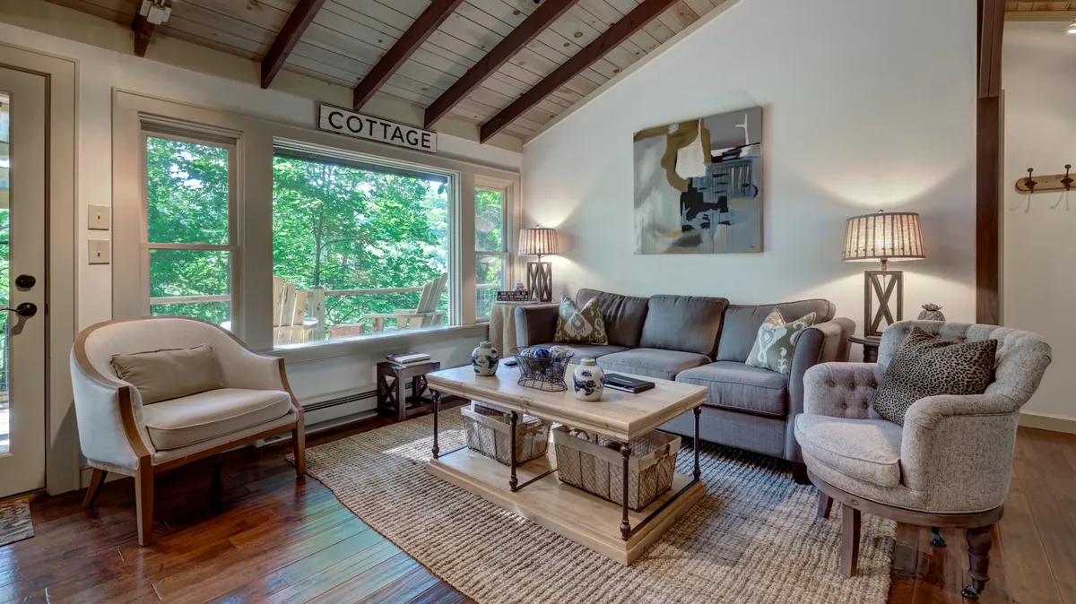 A bright, inviting living room with large windows framing tranquil views, plush sofa and armchairs gathered around a stylish coffee table, softly illuminated by a warm lamp. Thoughtful touches like decorative paintings and quality furnishings create a modern, comfortable ambiance perfect for relaxation and conversation.
