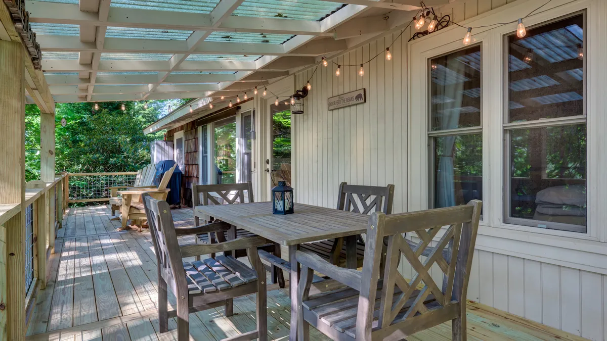 A beautifully set wooden dining table sits beneath glowing string lights and lanterns on a covered deck, surrounded by lush green foliage. Ample outdoor seating invites guests to linger over meals, while the inviting atmosphere suggests both daytime brunches and magical evenings.