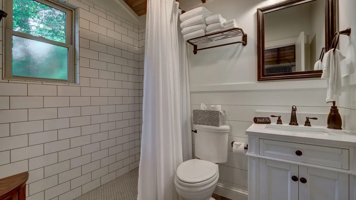 A modern en-suite bathroom with a vanity sink, framed mirror, shower area, and fresh towels, illuminated by natural light through a window overlooking greenery. The atmosphere is clean, inviting, and spa-like.
