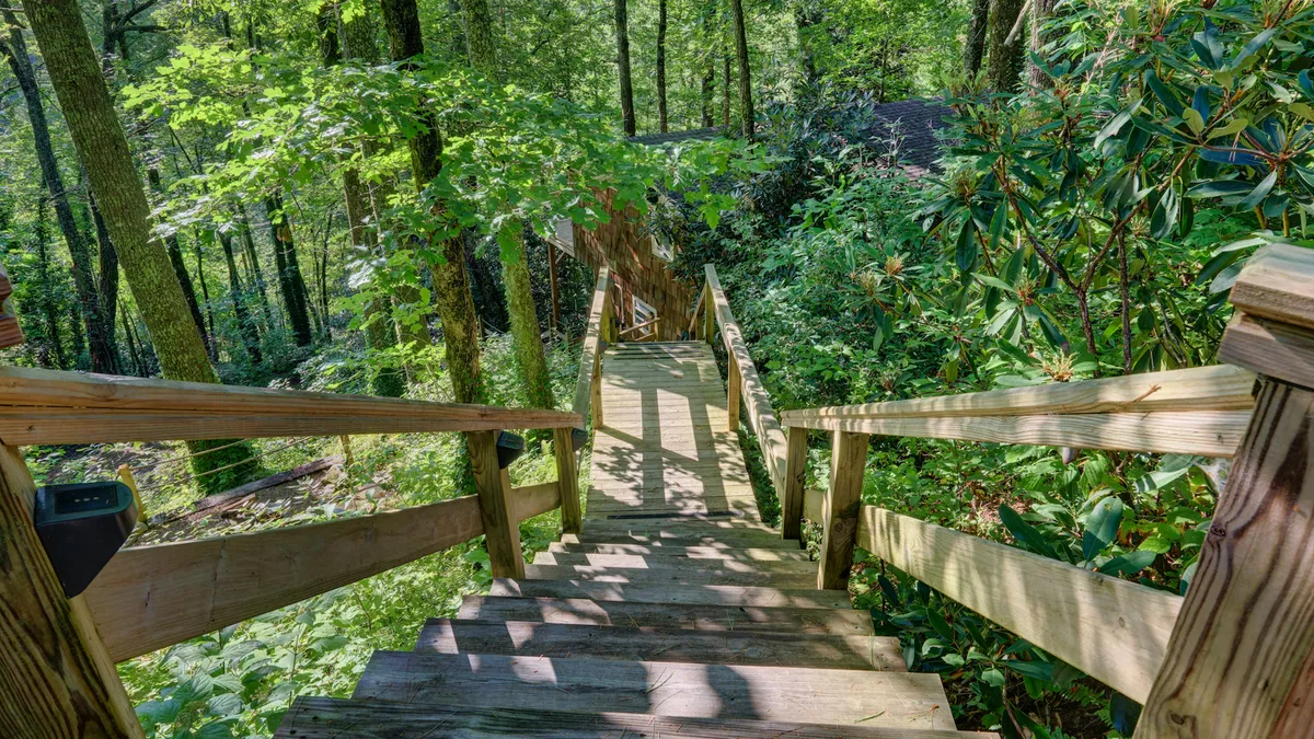 A wooden staircase winds through abundant greenery, dappled with sunlight filtering through the trees. The trail-like setting hints at nearby nature walks and the easy opportunity to discover wildlife, enjoy a picnic, or simply lose yourself in the tranquil outdoors.