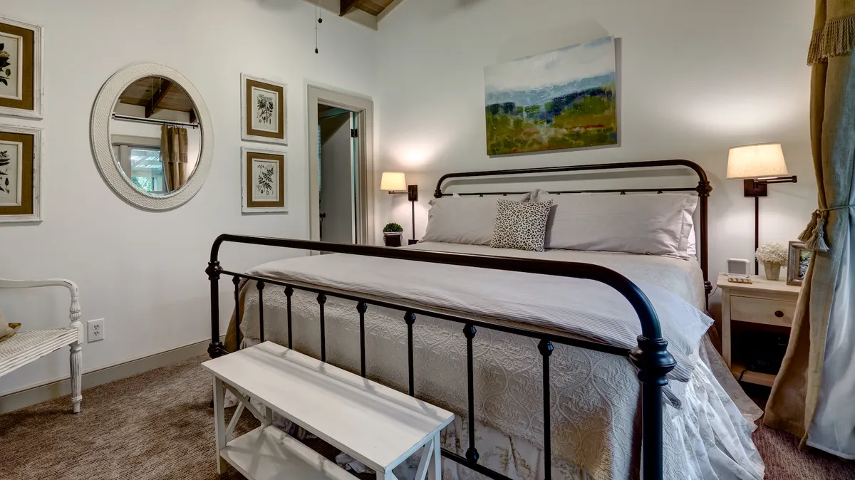 This image displays a serene king bedroom featuring an iron bed frame adorned with plush linens, flanked by stylish nightstands with lamps, and accented by tasteful artwork and a decorative mirror. Natural light and subtle décor create a calm, restful atmosphere.
