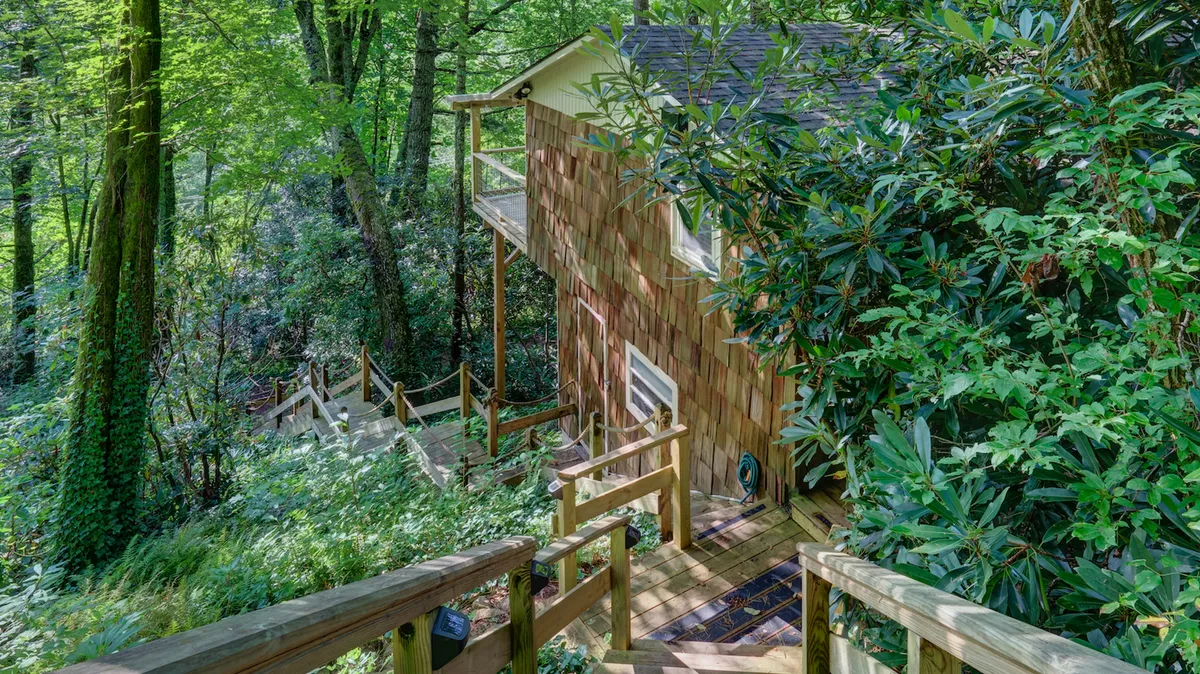 This image captures the wooden stairs leading down from the home's deck, surrounded by lush greenery and with the wooden exterior of the house peeking through the trees. The scene is bathed in soft, natural light, evoking a sense of calm and inviting viewers to imagine themselves stepping out into nature.